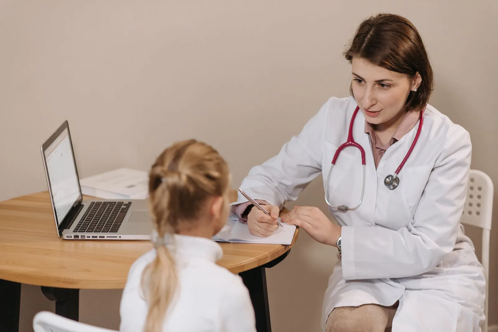 A clinician listening to a young patient while taking notes beside a laptop.